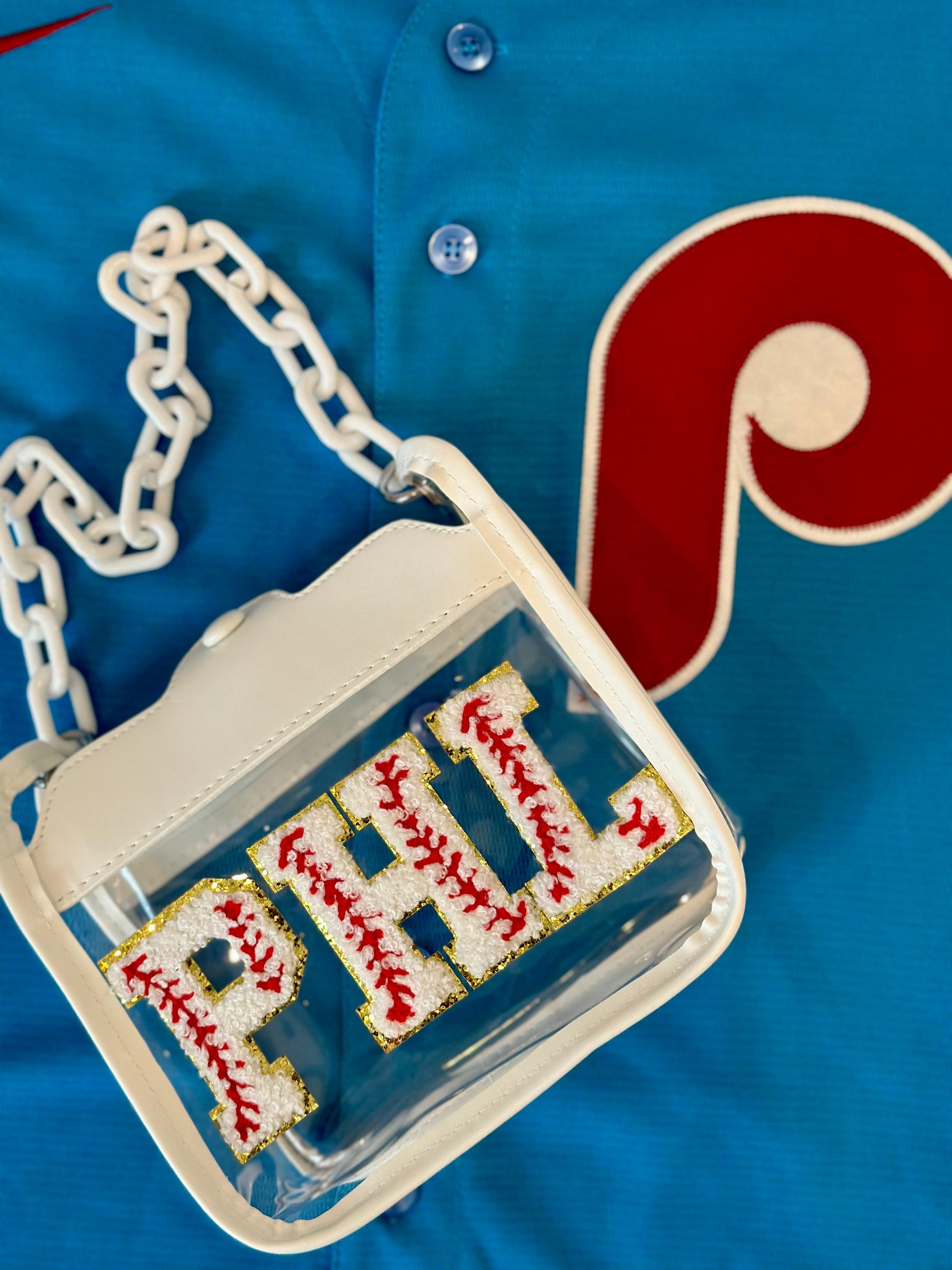 May include: A white clear plastic crossbody bag with a white chain strap. The bag has a white background with the word 'PHL' in red and gold letters with a baseball stitch design.