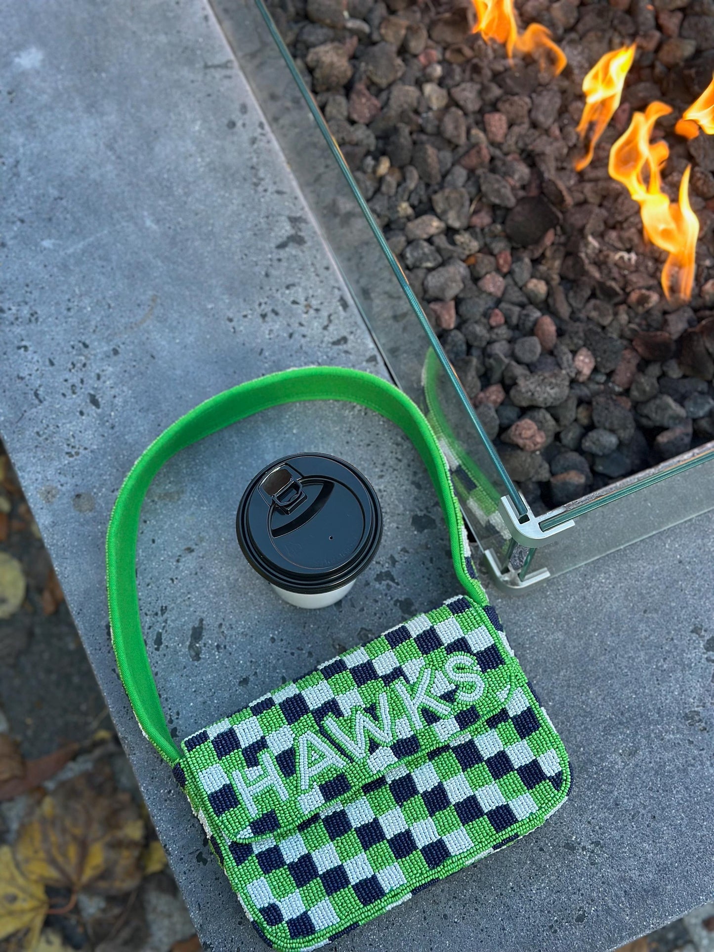 May include: A beaded handbag with a green handle and the word "HAWKS" in white letters. The bag features a checkerboard pattern in green, white, and navy blue. A disposable coffee cup with a black lid sits nearby.
