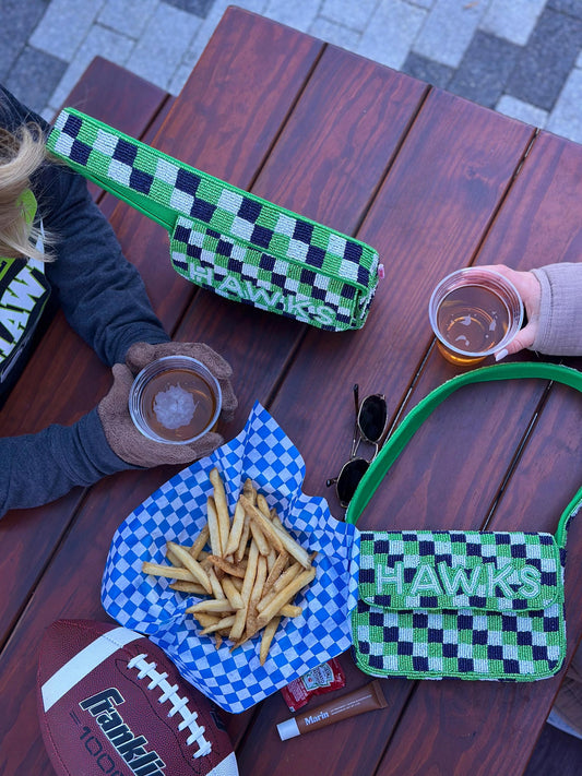 May include: Two beaded handbags with the word "HAWKS" in green and white. One is a long, rectangular clutch, and the other is a small shoulder bag. A football, fries, and drinks are on a wooden table.